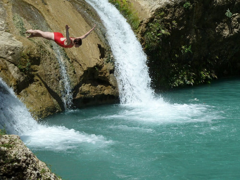 Woman jumping into the water next to the waterfall.